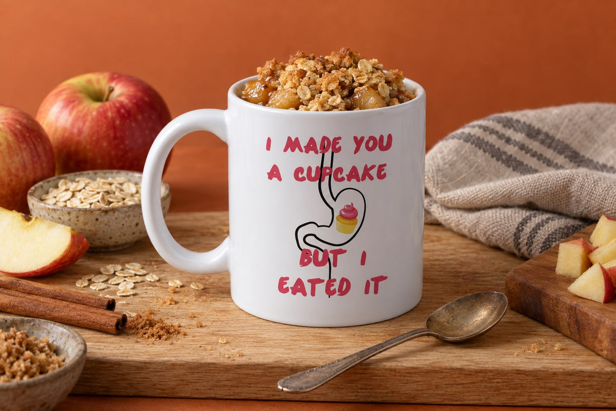 An apple crumble in the 'I Made A Cupcake But I Eated It' mug, with a loose golden crumble topping and apple juices bubbling at the edges, steam rising. A teaspoon lies on a linen tea towel beside it,